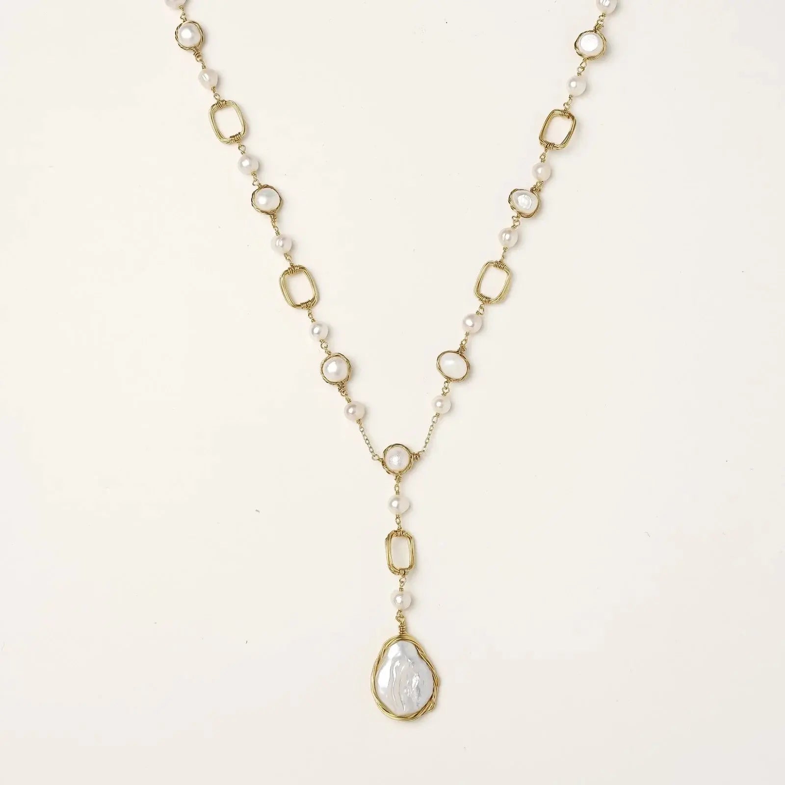 Y-shaped gold necklace with alternating geometric links, freshwater pearls, and baroque pearl pendant on cream background