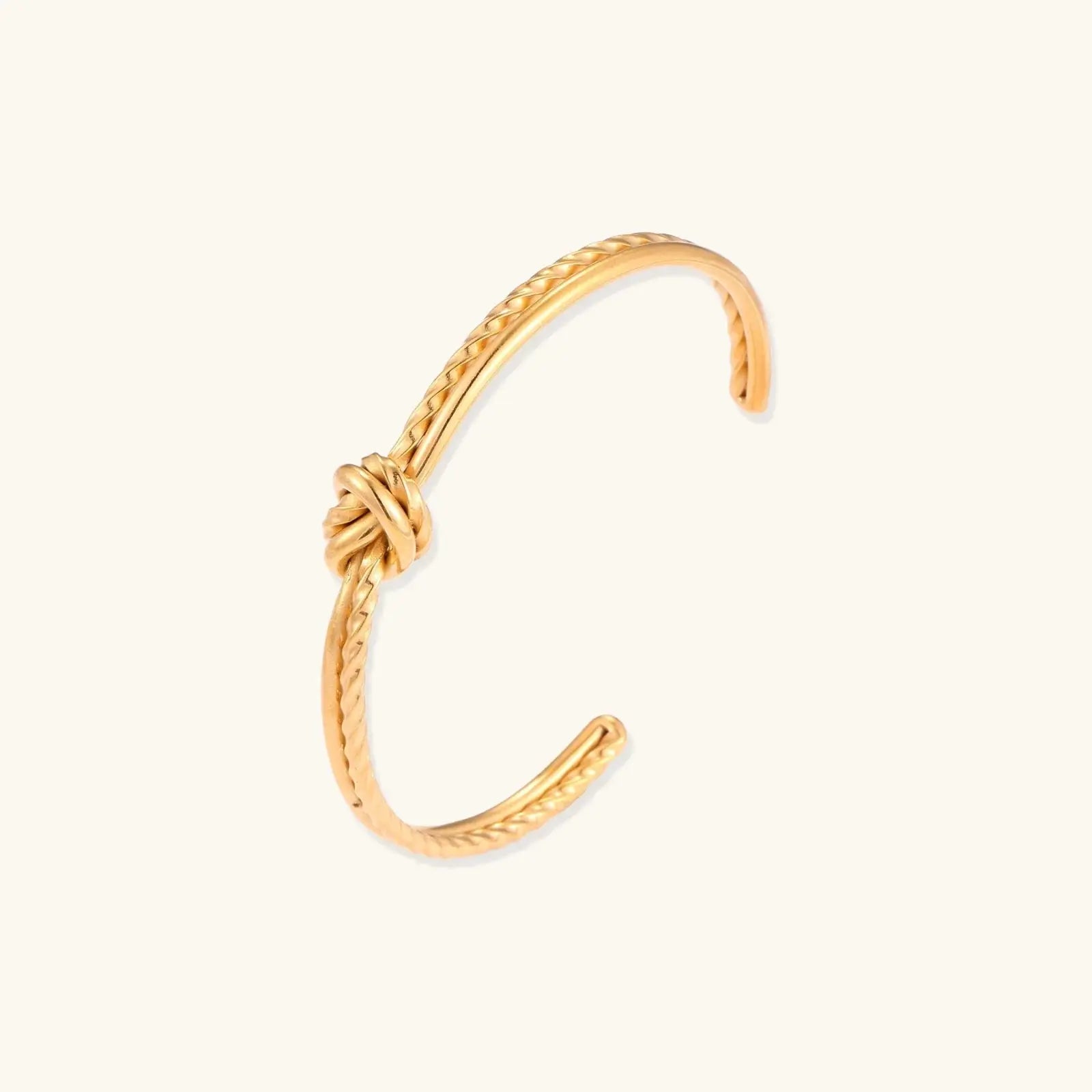 Gold cuff bracelet with twisted rope texture and decorative knot centerpiece on cream background.