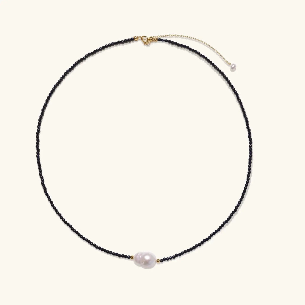 Delicate necklace with black blue sandstone beads and baroque pearl center on white background with gold accents.