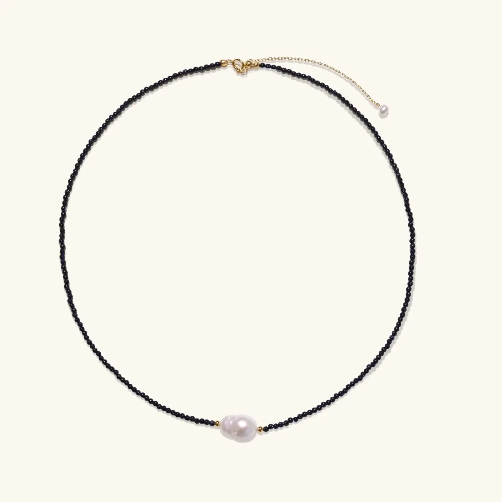Delicate necklace with black blue sandstone beads and baroque pearl center on white background with gold accents.